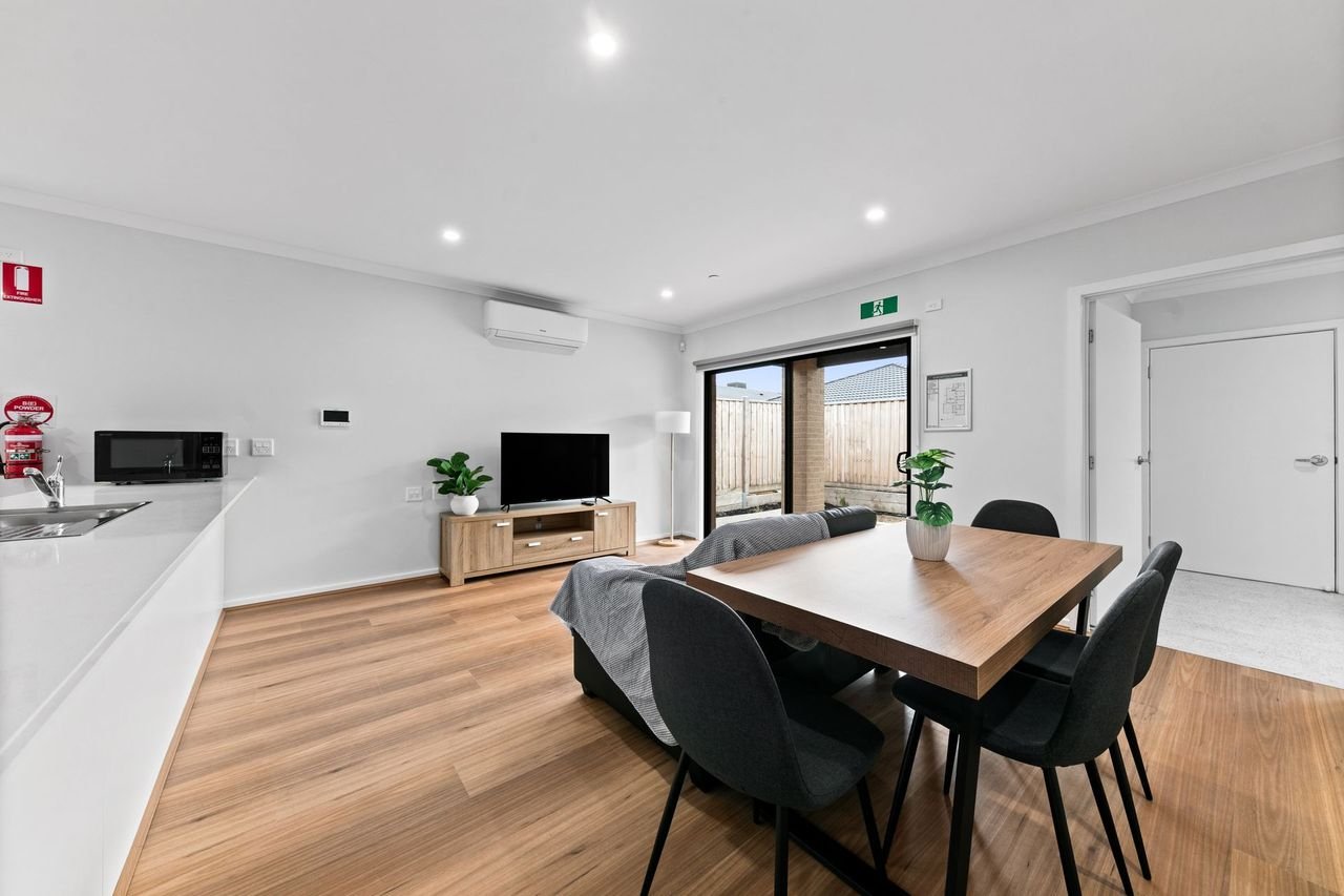 The open-plan kitchen, living and dining area of a Necessity home: a kitchen bench along one wall, a sofa facing a television, a dining table for four, and sliding doors opening onto a private fenced yard.